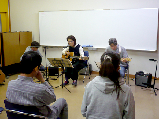 50代女性の生徒さんと講師のギター演奏とそれを聴く生徒さんたちの様子