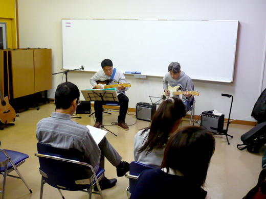 50代男性の生徒さんと講師のギター演奏とそれを聴く生徒さんたちの様子