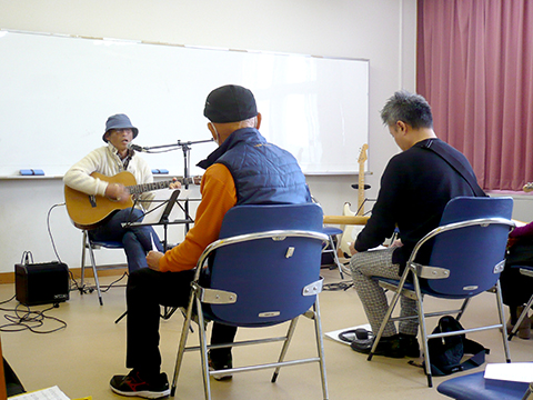 アコースティックギターを弾き語りする男性と演奏を聴く生徒さんの様子