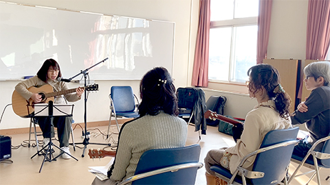 アコースティックギターを弾き語りする女性と演奏を聴く生徒さんの様子